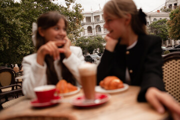 Two beautiful young girls are sitting in a French cafe, eating a croissant and drinking coffee with whipped cream. Teenagers are dressed in business style, black and white jacket in Old Money style