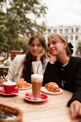 Two beautiful young girls are sitting in a French cafe, eating a croissant and drinking coffee with whipped cream. Teenagers are dressed in business style, black and white jacket in Old Money style
