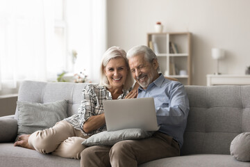 Smiling aged woman her hoary husband resting on sofa with laptop, watching movie via digital streaming services platform, enjoy electronic shopping using marketplace website. Leisure, technology usage