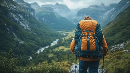 A hiker with a large backpack stands on a mountain trail, overlooking a lush green valley surrounded by towering cliffs under a cloudy sky.

