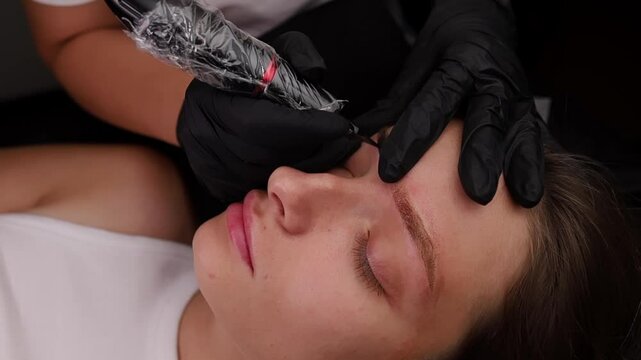 Young girl who lies with closed eyes master applies permanent makeup using machine technique on the eyebrows Light permanent eyebrow makeup, performing eyebrow tattooing