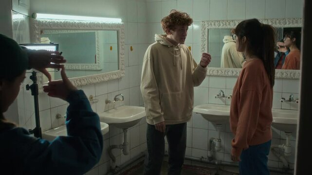 Full medium shot of two diverse female and male actors rehearsing quarrelling scene in washroom and director watching them through hands, giving advice before shooting
