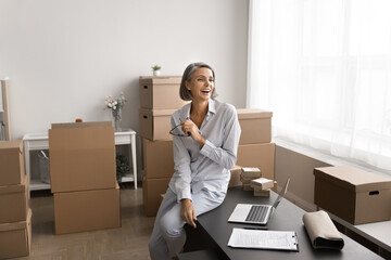 Mature woman sitting on desk surrounded by cardboard boxes, laughing, looking away. Warehouse...