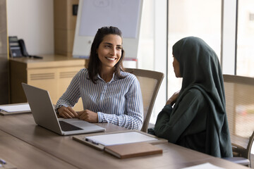 Happy young Latin business woman meeting and talking to Arab female colleague in hijab at workplace...