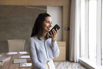 Happy successful business company owner woman speaking at mobile phone in office boardroom, keeping smartphone at open mouth, sending voice message, using online work communication