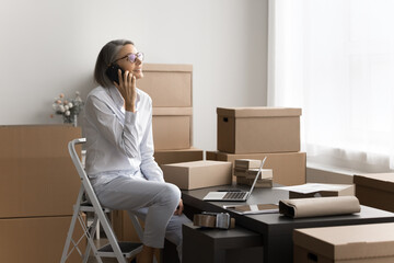 Mature 50s woman sitting on step stool, speaking on mobile phone, surrounded by cardboard boxes in home office setting, engaged in business conversation, related to managing or organizing shipments
