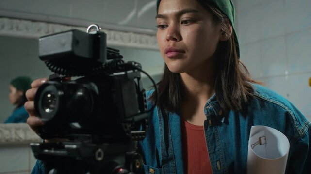 Chest up shot of young Caucasian female operator in denim jacket and cap setting up professional video camera before shooting in movie set, changing focus