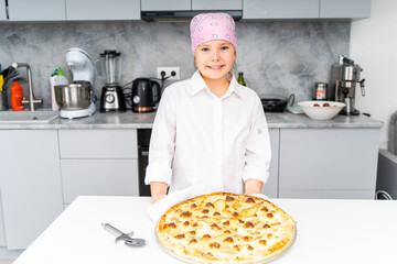 A little teenage girl in a chef's jacket shows her pizza with cheese and pear against the background of a gray kitchen