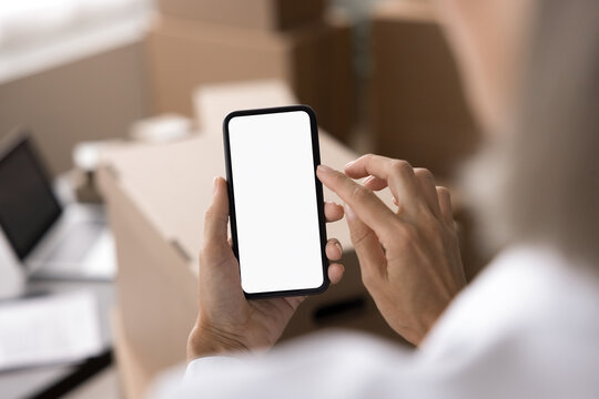 Unknown woman, small business owner, holding smart phone with white mock up screen, close up view over female shoulder. Organizing, scheduling, mobile apps, ordering delivery courier services usage