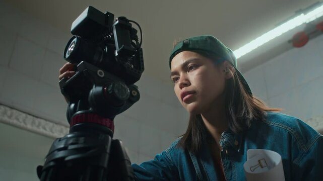 Low angle arc shot of young Caucasian female camera operator in denim jacket and cap setting up filmmaking equipment and getting ready for movie shooting in film set