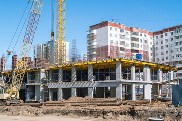 A construction site where tower and crawler cranes perform lifting work. A new house is being built on the site of the old houses. A road is being paved near the house under construction.