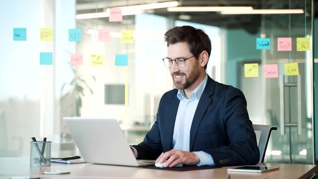 Confident businessman smiles while working on laptop at workplace in business office. Businessman engaged in tasks related to workplace productivity. Handsome worker in formal suit works on a computer
