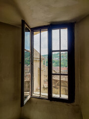 Open window in old building, ancient fortress. View through grid window overlooking green landscape and distant hills. Sunlight streams through mesh, creating serene atmosphere.