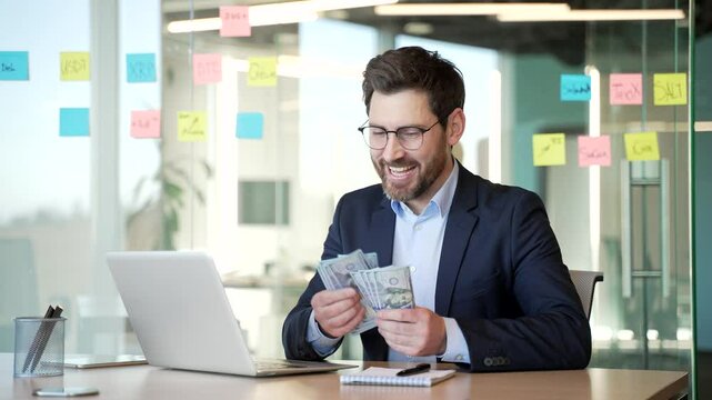 Happy businessman in suit counting dollar bills and pointing finger at laptop screen sitting in a business office. Handsome male is satisfied with earning money using an application on his computer