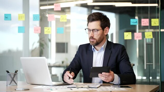 Busy businessman working doing paperwork sitting at desk at workplace in business office. Confident financier in a formal suit calculates on a calculator, takes notes, types on laptop computer