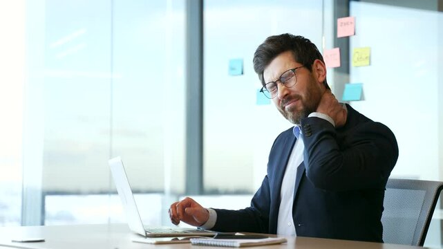 Tired businessman in uit suffering from neck pain while working on laptop sitting at desk at workplace in business office. Upset male worker feels discomfort, massages and rubs sore muscles, stretches