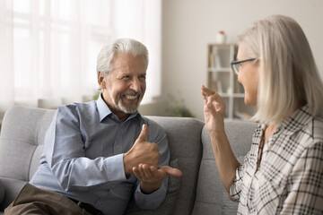 Older hoary husband sit on sofa at home communicates with mature wife using gestures. Older man with hearing loss, physical disability interact with smiling positive spouse showing each other symbols