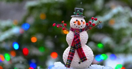 A small wax snowman figurine with black Top Hat and Red Scarf is outside while it is snowing and having a Christmas Tree with Lights Blinking in the Background. - Powered by Adobe