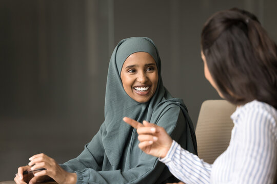 Cheerful beautiful young Arabian and European business women talking at office table, chatting on work break, smiling, laughing, discussing project, office life, enjoying corporate diverse friendship