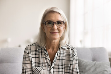 Head shot portrait attractive mature woman in glasses sit on sofa looking at camera, take part in...