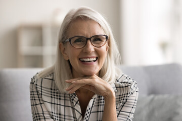 Close up head shot portrait cheerful confident mature 60s woman posing for camera, sit on sofa at home, having white perfect teeth advertising professional dental services. Profile picture, front view