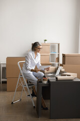 Woman sit on step ladder in room filled with cardboard boxes, hold sheet of paper, look at laptop on desk, engaged in workflow related to shipping or packaging products, control e-commerce operations