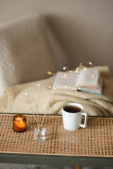 Cup of coffee with candle and home perfume in glass bottle over knitted sweater on accent chair close up. Winter holiday season.