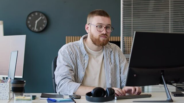 Medium portrait of young bearded Caucasian man wearing smart casual clothes looking at camera while working on computer