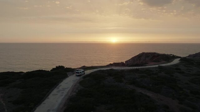 Aerial view of a white camper driving on the coastal road by the calm sea at golden sunset. Roadtrip traveling and driving by the seaside in the evening. Vacations on the road. 