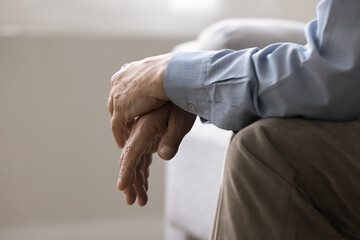 Cropped view, folded hands of unknown senior, man sitting on couch indoors. Close up shot of...