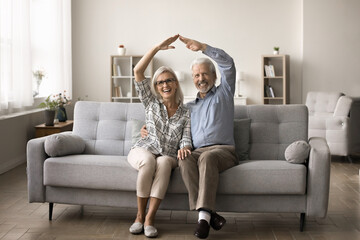 Happy senior homeowners couple sit on sofa in modern living room, joining their hands showing roof...