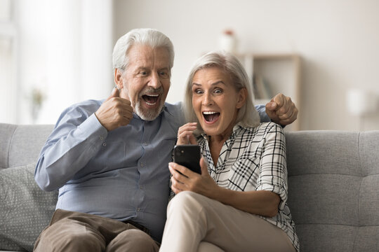 Overjoyed mature spouses sit on sofa read sms on smartphone, clenched their fists making yes gesture celebrate jackpot, bet, gambling win. Ger sell-out offer, bank loan approval, relish victory moment