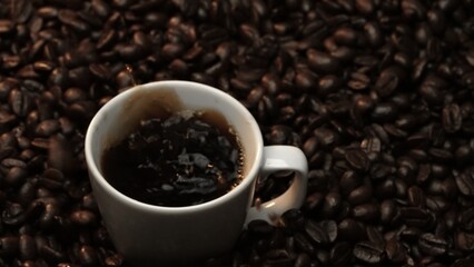 Fototapeta premium Close up of coffee bean drop in to hot espresso with pile of coffee seed. Macro of freshly roasted bean falling in to cup with aromatic smoke. Slow motion. Organic coffee seed concept. Comestible.