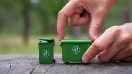 Hand pours paper and glass waste from toy green bins set on a tree stump in the forest, then sorts and places it back into the recycling bins.