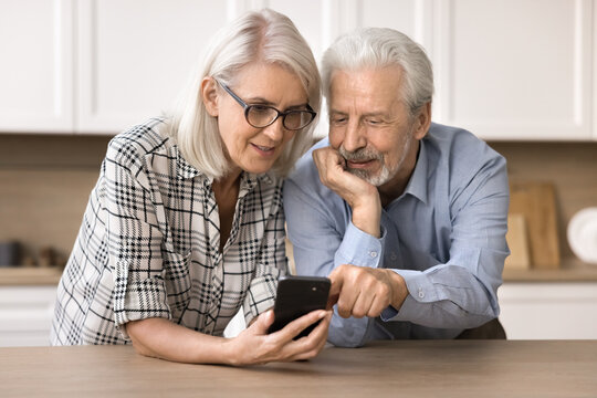 Interested mature spouses learn received message, read commercial offer in sms, use smart phone standing at kitchen table, ordering products via on-line supermarket app, review new mobile application