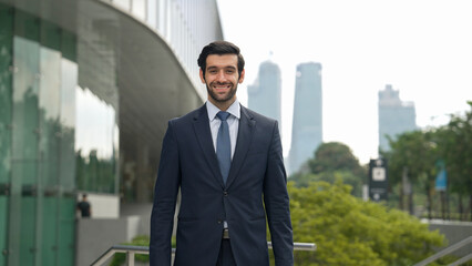 Professional businessman standing at city or park. Smiling project manager looking at camera while smiling arms with confident. Closeup of investor wearing suit at architectural building. Exultant.