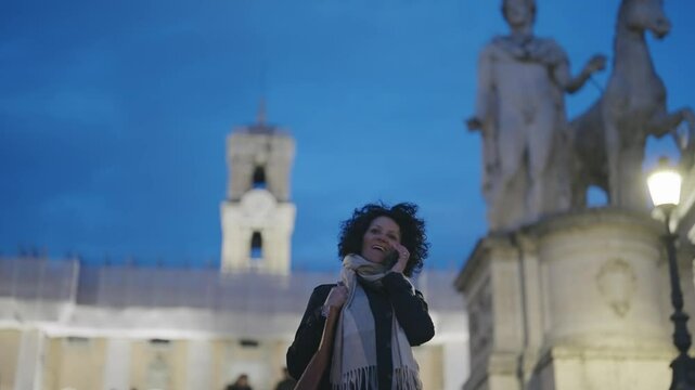 A businesswoman enjoys a phone conversation with defocused background of the Cordonata Capitolina in Rome during twilight, with historic statues and architecture in the background.