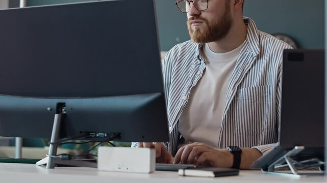 Medium shot of young Caucasian sysadmin with beard on face typing on desktop computer in IT company office