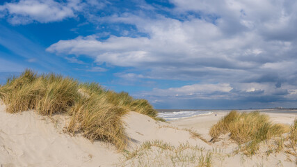 Golden dunes topped with grass overlook a tranquil beach, with waves gently kissing the Zandmotor shore near Kijkduin under a partly cloudy sky. In the distance, two people can be seen walking.