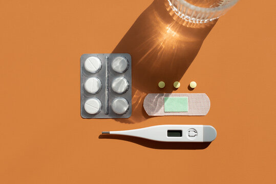 Minimalist collection of medical items with glass of water on orange background. White digital thermometer, adhesive bandage, round yellow pills and blister pack of white tablets