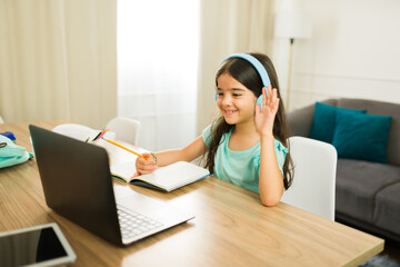 Happy Hispanic girl with headphones waving at laptop screen while studying at desk in living room