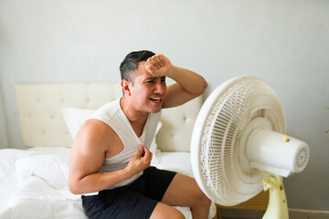 Sweaty man suffering from summer heat, feeling hot and exhausted in his bedroom while sitting on...