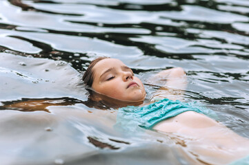 Obraz premium Girl child swims on back, rests lying in water on lake, sea in summer. Photography, portrait, sport concept, childhood.