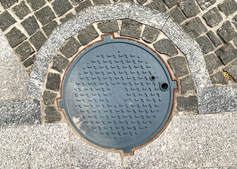 Sewer black round cast iron manhole cover embedded in a cobblestone pavement, showcasing a geometric pattern and weathered texture. Top view. Close up