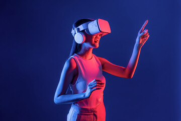 Smart female stand with surrounded by cyberpunk neon light wear VR headset connecting metaverse, futuristic cyberspace community technology. Woman using finger pointing virtual object. Hallucination.