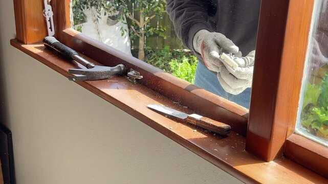 Close up of a glazier's gloved hands working with a metal scraper to smooth a wooden window frame surface in preparation for installation of a new glass pane in New Zealand