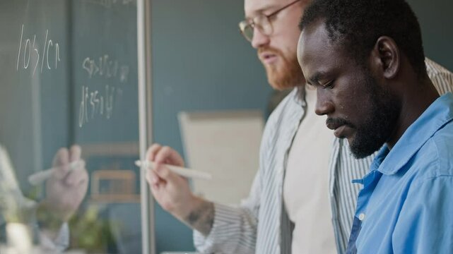 Young Black male programmer holding digital tablet in hands, his Caucasian colleague writing code on glass wall