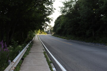 A road and an hill in the woods. A sidewalk beside a road and in summer. The asphalt on a road and transportation. 