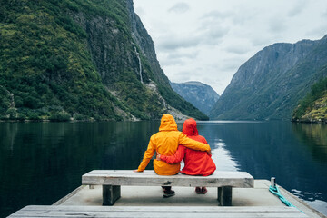 Happy couple dressed in bright windproof jackets sitting and hugging on bench on the Norwegian Nærøyfjord bank in Gudvangen, Norway. Traveling, beauty in Nature and relations concept.