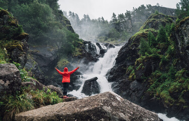 Woman trekker dressed bright red waterproof jacket with enjoying Låtefossen Waterfall, Norway